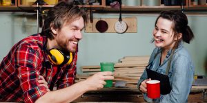 Man and girl carpenters having break from indoor work in wood workshop. Coffee time, drink, share experiences, discuss. Teacher and student. Laughing smile.